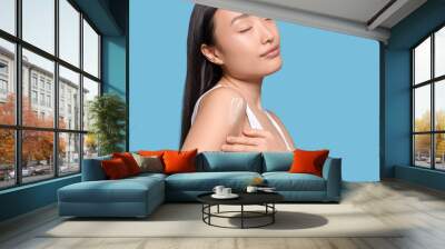 Beautiful young Asian woman applying body cream onto shoulder on light blue background, space for text Wall mural