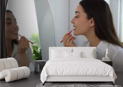 Beautiful young woman applying cosmetic pencil on lips near mirror indoors Wall mural