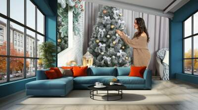 Beautiful young woman decorating Christmas tree at home Wall mural