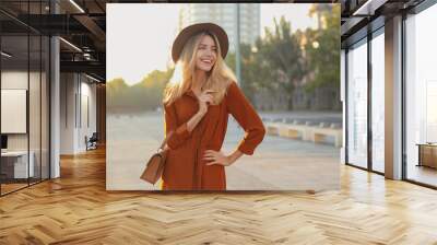 Beautiful young woman in stylish red dress and hat with handbag on city street Wall mural