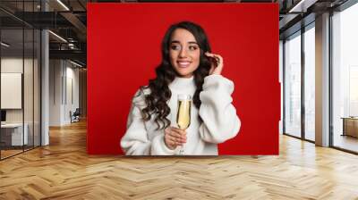 Beautiful young woman in warm sweater holding glass of champagne on red background. Christmas party Wall mural