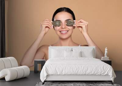 Beautiful young woman putting green tea bags on skin around eyes against beige background Wall mural