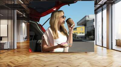 Beautiful young woman with doughnut drinking coffee near car at gas station Wall mural