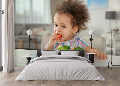 Cute African-American girl eating vegetables at table in living room Wall mural