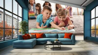 Cute kids reading book on floor while other children playing together in kindergarten Wall mural