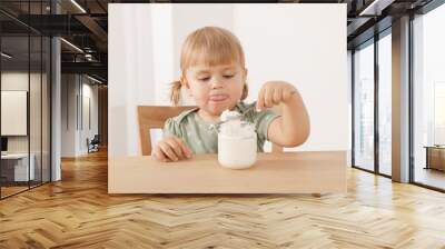 Cute little child eating tasty yogurt with spoon at wooden table indoors Wall mural