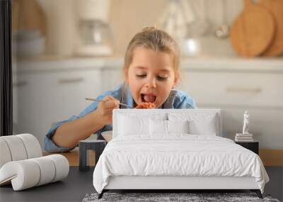 Cute little girl eating tasty pasta at table in kitchen Wall mural