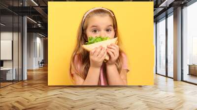 Cute little girl eating tasty sandwich on yellow background Wall mural