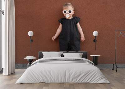 Cute little girl wearing stylish clothes with sunglasses and pacifier near brown wall outdoors Wall mural