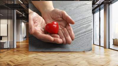 Elderly woman holding red heart in hands at grey wooden table, closeup Wall mural