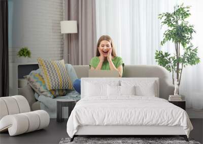 Emotional young woman with laptop celebrating victory on sofa at home Wall mural