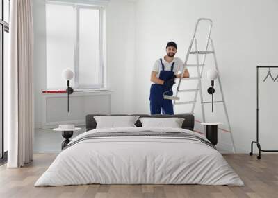 Full length portrait of handyman with roller brush near ladder indoors, space for text. Professional construction tools Wall mural