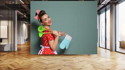 Funny young housewife with oven mitten and cooking utensils on color background Wall mural