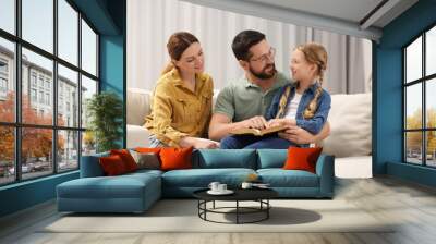 Girl and her godparents reading Bible together on sofa at home Wall mural