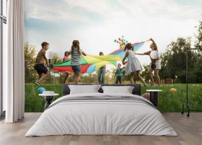 Group of children and teachers playing with rainbow playground parachute on green grass, low angle view. Summer camp activity Wall mural