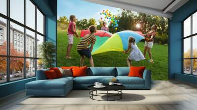 Group of children and teachers playing with rainbow playground parachute on green grass. Summer camp activity Wall mural