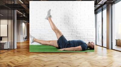 Handsome young man in sportswear doing scissors exercise on artificial green lawn near brick wall Wall mural