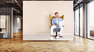 Handsome young man with laptop in armchair near color wall indoors. Space for text Wall mural