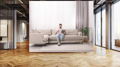 Happy man reading book on sofa near window with beautiful curtains in living room Wall mural