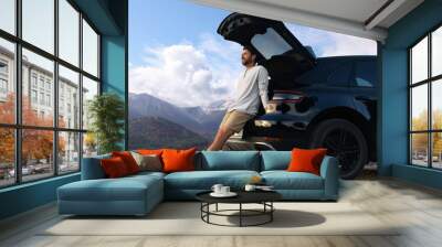 Happy man sitting in trunk of modern car in mountains Wall mural