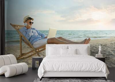 Happy man with laptop talking by mobile phone on beach. Business trip Wall mural