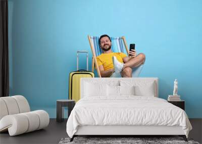 Happy man with smartphone sitting on folding chair and suitcase against light blue background Wall mural