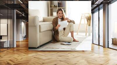 Happy woman reading letter while sitting on floor near sofa at home Wall mural