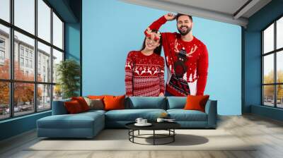 Happy young couple in Christmas sweaters, reindeer headband and funny glasses on light blue background Wall mural