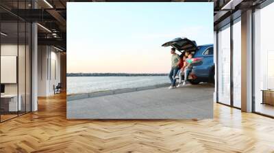 Happy young family sitting in car trunk on riverside Wall mural