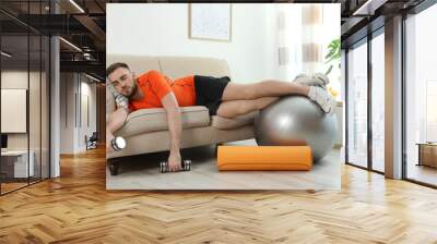 Lazy young man with sport equipment on sofa at home Wall mural