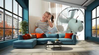 Little girl enjoying air flow from fan on sofa in living room. Summer heat Wall mural