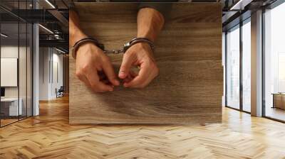 Man in metal handcuffs at wooden table, closeup. Space for text Wall mural