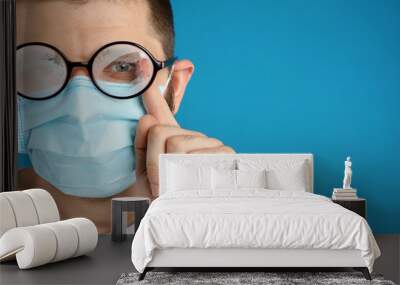 Man wiping foggy glasses caused by wearing disposable mask on blue background, space for text. Protective measure during coronavirus pandemic Wall mural