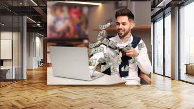 Man with modern laptop and flying dollar banknotes at table indoors. People make money online Wall mural