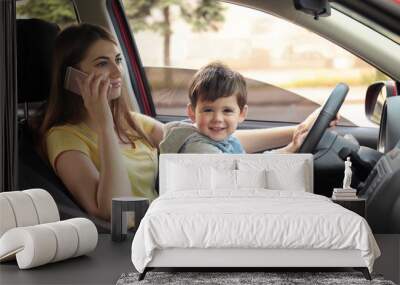 Mother with little son on knees driving car and talking by phone. Child in danger Wall mural