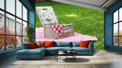 One picnic wicker basket with checkered napkin, tableware and blanket on green grass. Space for text Wall mural