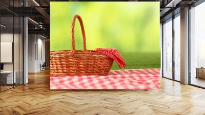 One picnic wicker basket with checkered napkin and blanket on grass against blurred green background. Space for text Wall mural