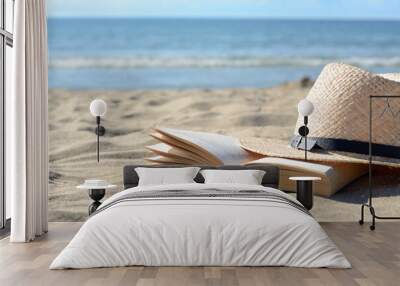 Open book and straw hat on sandy beach near sea, space for text Wall mural