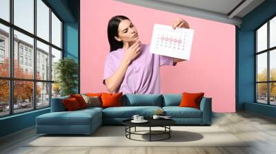 Pensive young woman holding calendar with marked menstrual cycle days on pink background Wall mural