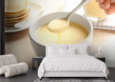 Person holding spoon with condensed milk over bowl on table, closeup. Dairy products Wall mural