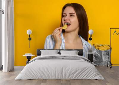 Pretty young woman eating tasty potato chips on yellow background Wall mural