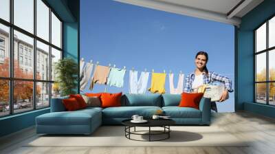 Smiling woman holding basket with baby clothes near washing line for drying against blue sky outdoors Wall mural