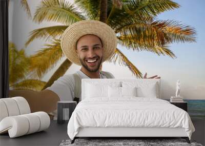 Smiling young man in straw hat taking selfie at beach Wall mural