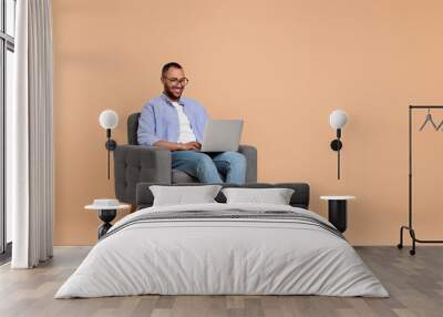 Smiling young man working with laptop in armchair on beige background, space for text Wall mural