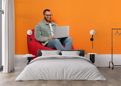 Smiling young man working with laptop on beanbag chair near orange wall, space for text Wall mural