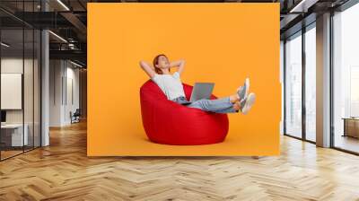 Smiling young woman with laptop sitting on beanbag chair against yellow background Wall mural