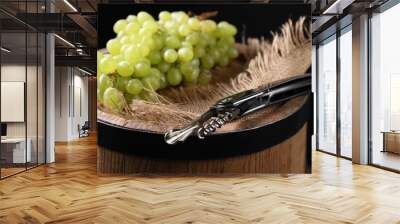 Sommelier knife with corkscrew and grapes on wooden barrel against black background, closeup Wall mural