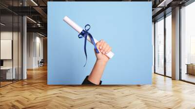 Student holding rolled diploma with ribbon on light blue background, closeup Wall mural