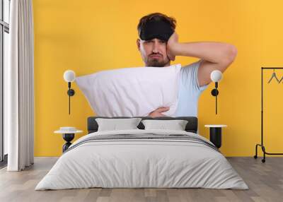 Unhappy man with pillow and sleep mask on yellow background. Insomnia problem Wall mural