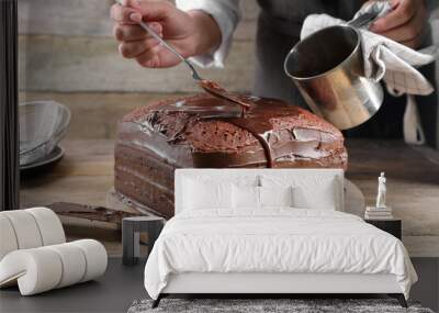 Woman pouring chocolate cream into homemade sponge cake at wooden table, closeup Wall mural